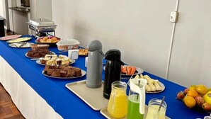A buffet table with various food items and beverages, including fruits, pastries, and drinks, set up in a room with a tiled floor and white walls. - Hotel Jaú (Jau)