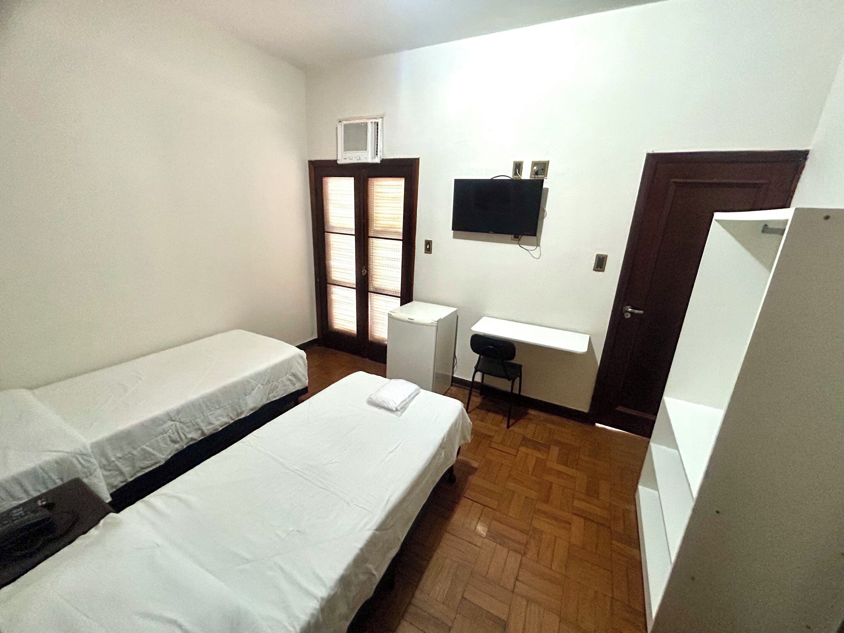 A small, clean room with two single beds, a desk, a chair, a TV mounted on the wall, and a door.