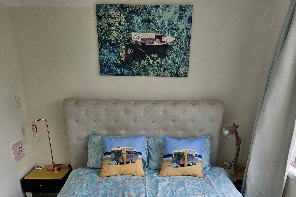 A bedroom with a large bed, a headboard, two bedside tables, a nightstand, a lamp, a window with curtains, and a painting of a boat on water.