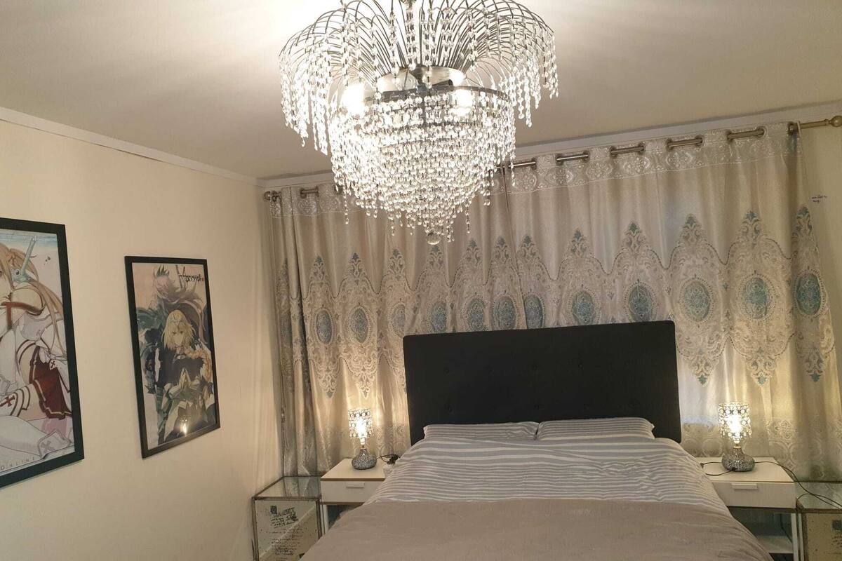 A bedroom with a large bed, a chandelier, patterned curtains, and framed artwork on the wall.