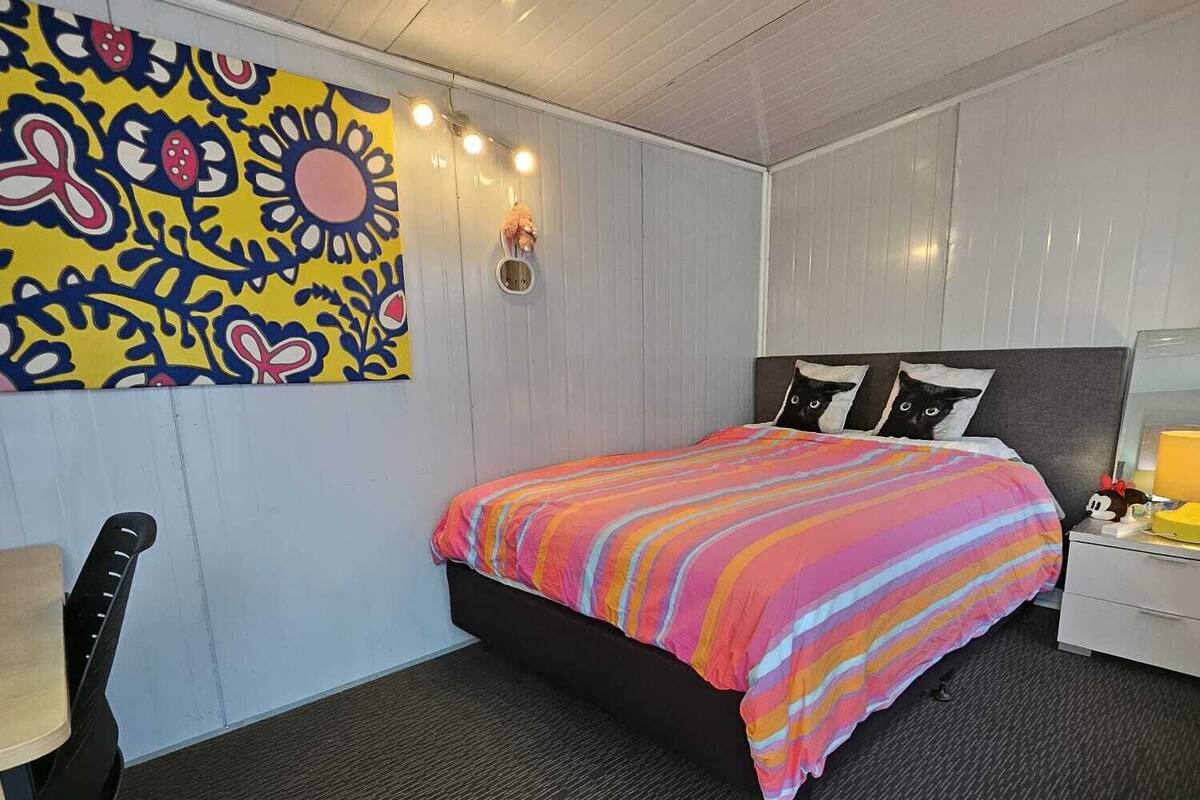 A bedroom with a bed, a desk, a chair, a wall with a colorful floral pattern, and a ceiling light.
