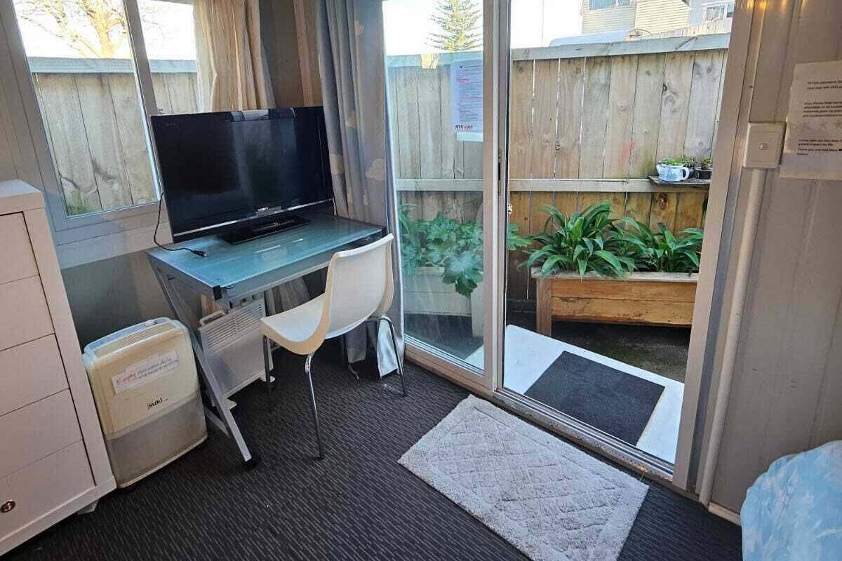 A compact room with a desk, chair, and a television. It features a sliding glass door leading to an outdoor area with a wooden fence and plants.