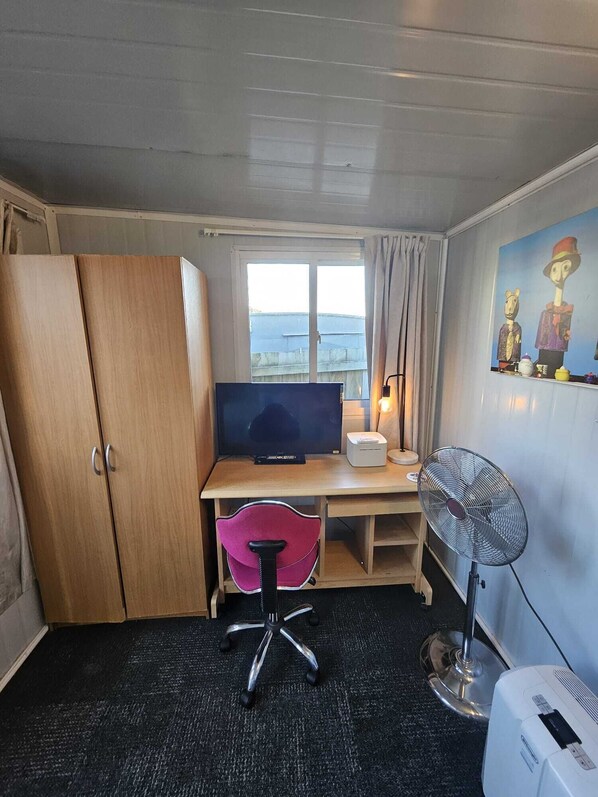 Habitación pequeña con escritorio de madera, televisor, silla de oficina rosa, ventilador de pie y un cuadro en la pared.