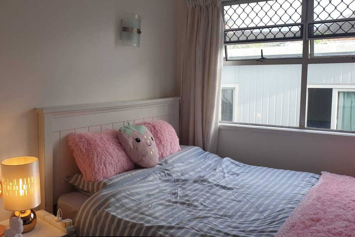 A bedroom with a bed, a nightstand with a lamp, and a window with a view of the outside.