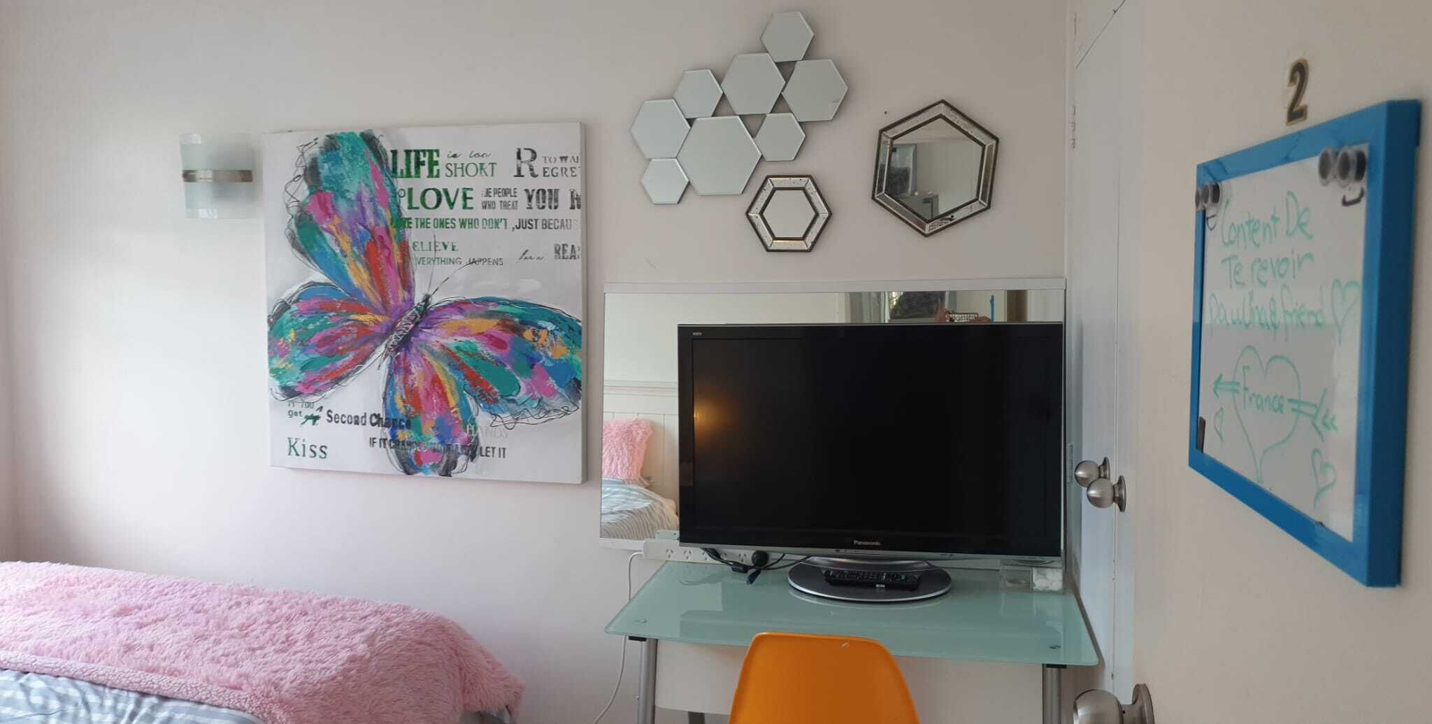 Ein Schlafzimmer mit einem Bett, einem Fernseher auf einem Glastisch, einem Schmetterlingsbild, sechseckiger Wanddekoration und einer Tür mit blauem Rahmen.