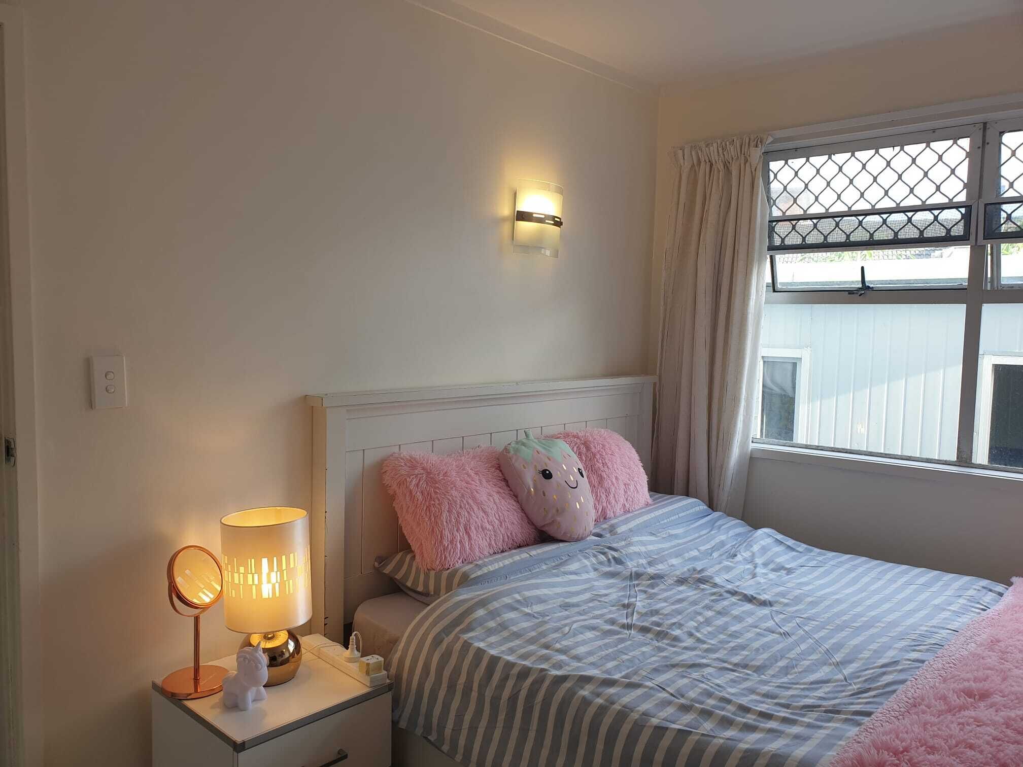 A bedroom with a bed, a nightstand with a lamp, a wall-mounted light, and a window with curtains.