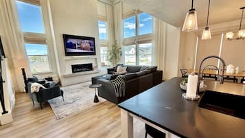 A modern living room with a large TV, comfortable seating, a fireplace, and a kitchen area with a sink and countertop.