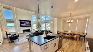 A modern kitchen with a central island, a dining area with a wooden table and chairs, a living room with a sofa and TV, and large windows allowing natural light. - Silicon Slopes Retreat (Lehi)