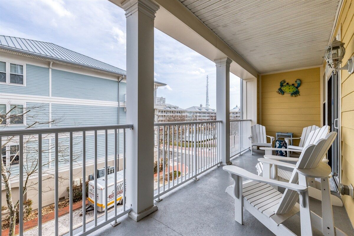 A covered porch with white Adirondack chairs, a small table, and a decorative frog wall art.