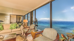 A spacious living area with a large window offering a view of the ocean. There is a sofa with tropical cushions, a round table with a lamp, and a decorative sculpture. - The Mahana at Kaanapali (Lahaina)
