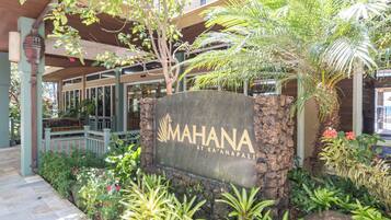A sign for 'MAHANA AT KA'ANAPALI' with a stone base, surrounded by lush greenery and a wooden pergola.