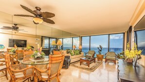 A spacious living area with a large glass dining table surrounded by wicker chairs, a sofa, and a coffee table. There are ceiling fans, a TV, and a view of the ocean through large windows. - The Mahana at Kaanapali (Lahaina)