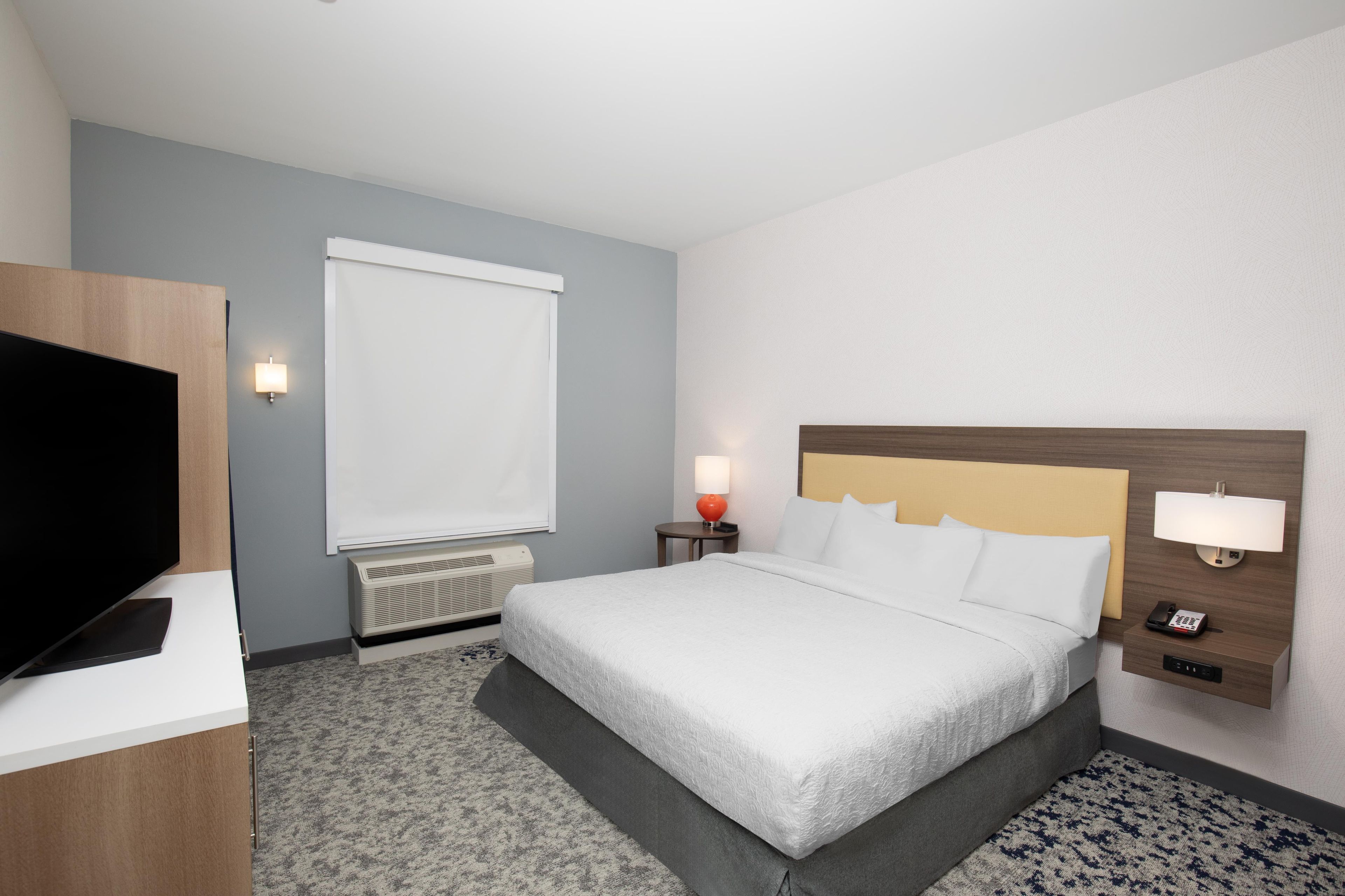 A hotel room with a bed, a television, a heater, a window with a blind, and a bedside lamp.