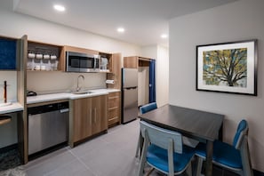 A compact kitchen with a dishwasher, microwave, and refrigerator. There is a dining table with chairs and a framed painting on the wall. - Home2 Suites By Hilton Hoover Birmingham (Birmingham)
