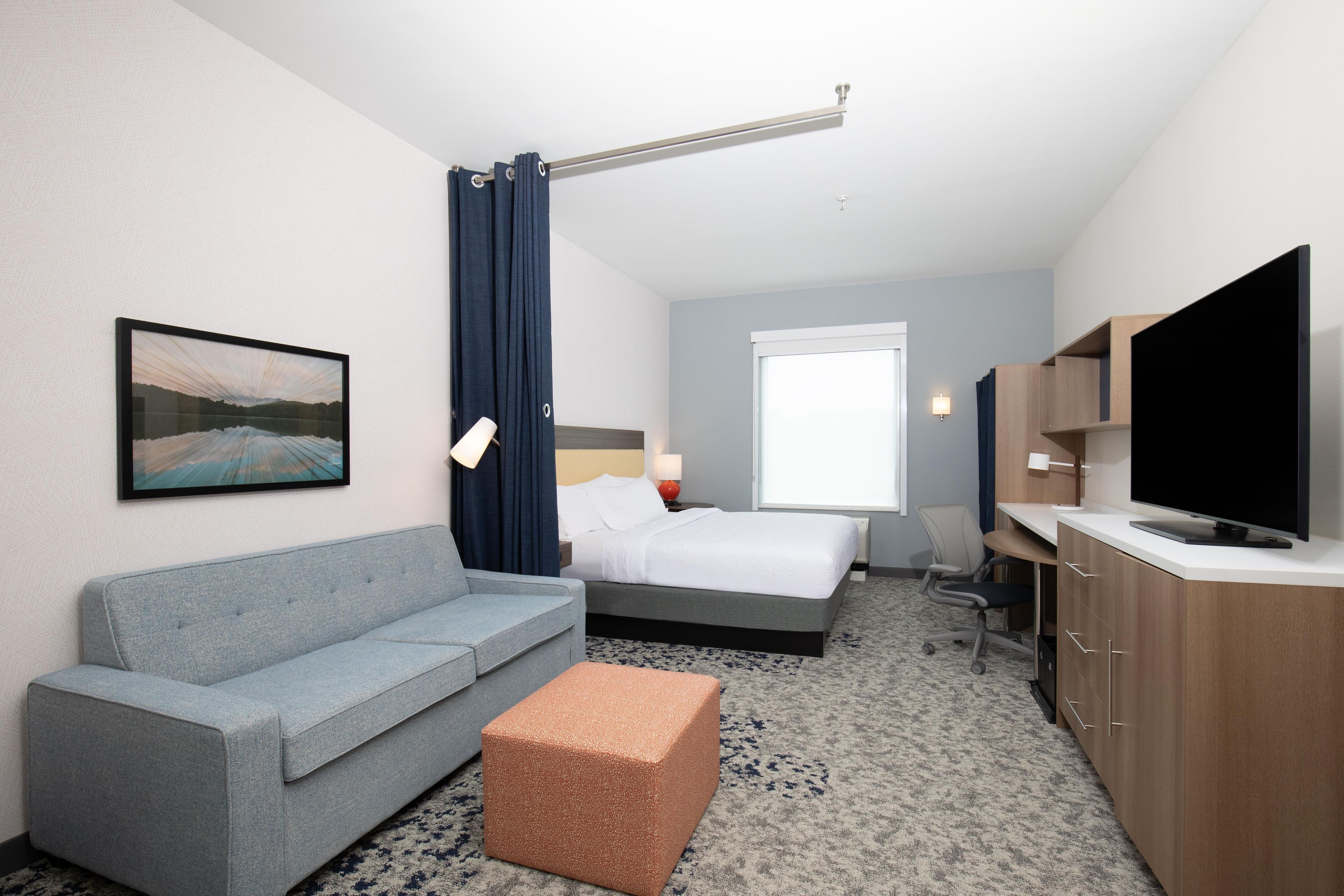 A hotel room with a grey sofa, a bed with white linens, a television, and a painting on the wall.