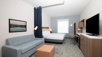 A hotel room with a grey sofa, a bed with white linens, a television, and a painting on the wall.