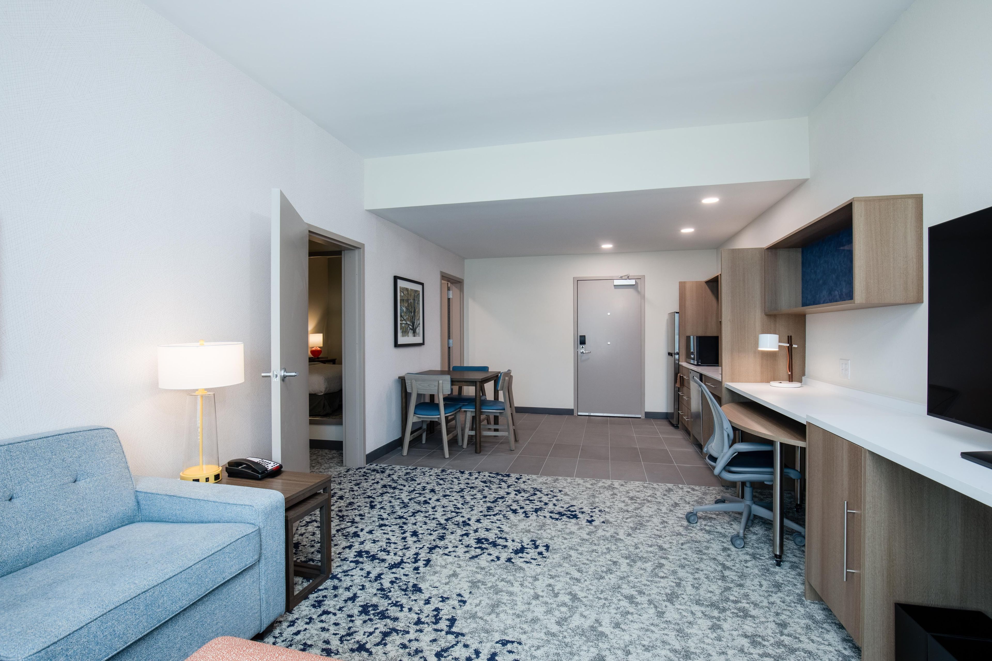A compact hotel room with a sofa, a desk, a chair, a small table, a lamp, a TV, and a door leading to a bathroom.