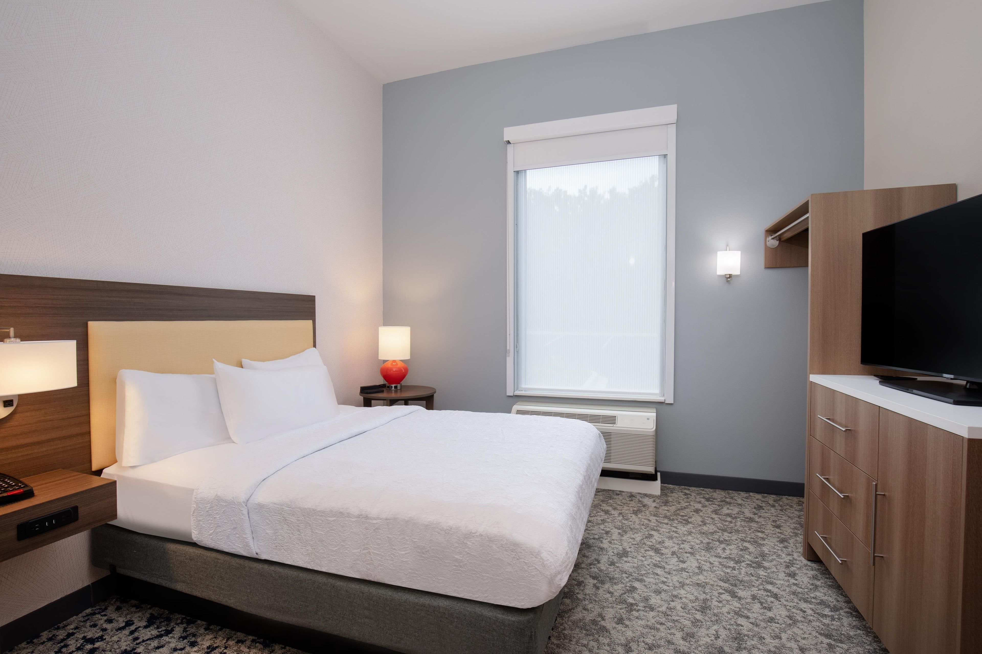 A hotel room with a bed, bedside table, lamp, TV, and a window with blinds.