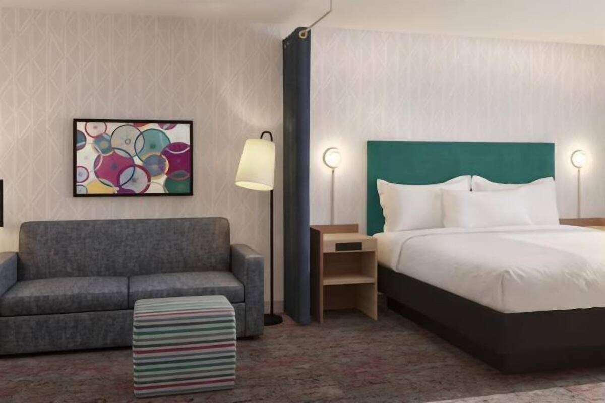 Een hotelkamer met een bed, een slaapbank, een nachtkastje, een lamp en een kleurrijk abstract schilderij aan de muur.