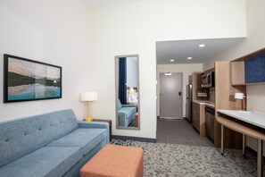 A modern hotel room with a sofa, a small ottoman, a painting on the wall, a mirror, a lamp, a kitchen area with a microwave, and a door leading to another room. - Home2 Suites By Hilton Hoover Birmingham (Birmingham)