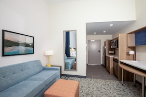 A modern hotel room with a sofa, a small ottoman, a painting on the wall, a mirror, a lamp, a kitchen area with a microwave, and a door leading to another room.