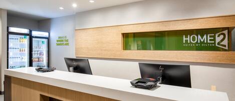 A reception desk with a sign reading 'HOME 2 SUITES BY HILTON' and a wall-mounted refrigerator with snacks.