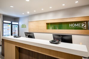 Een receptiebalie met een bordje met de tekst 'HOME 2 SUITES BY HILTON' en een in de muur bevestigde koelkast met snacks.