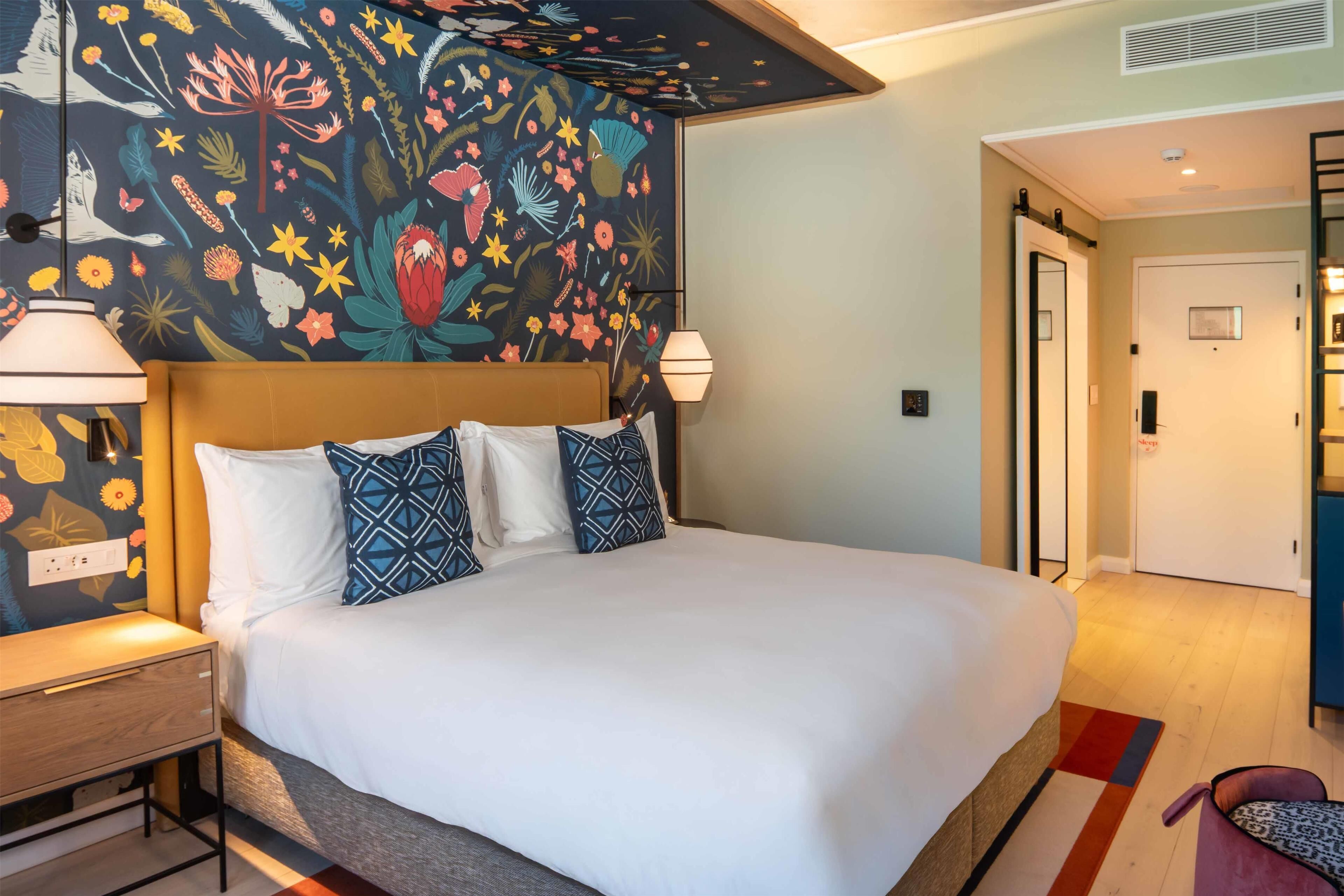 A hotel room with a large bed, a nightstand, a lamp, a wall with a floral mural, and a door leading to another room.