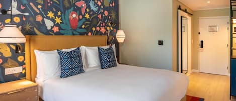 A hotel room with a large bed, a nightstand, a lamp, a wall with a floral mural, and a door leading to another room.