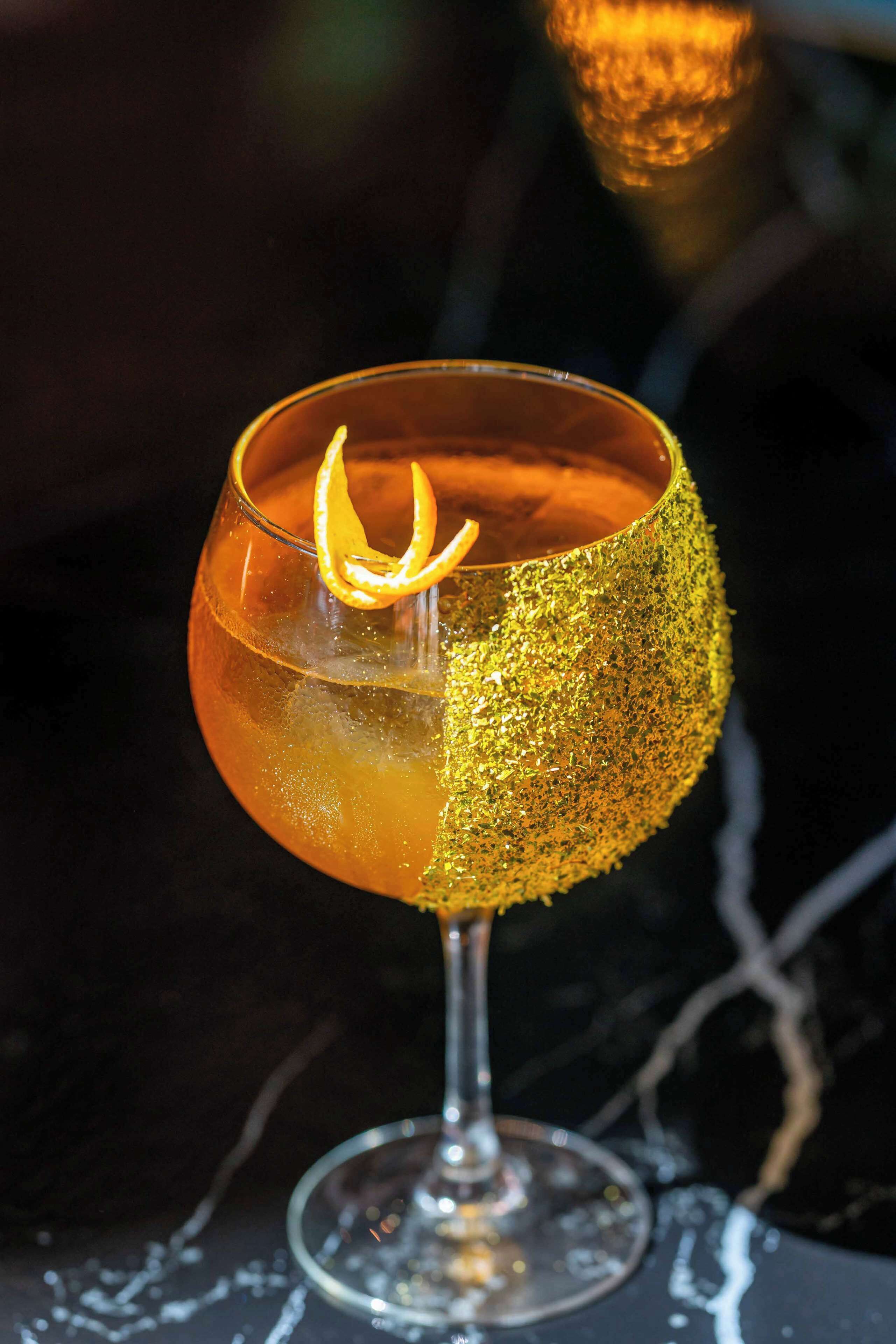 Cocktail-lasi, jonka reunassa on kultainen, kimalteleva koristelu ja sitruunaviipale koristeena.