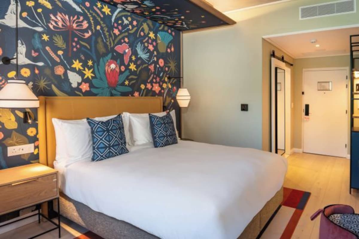 Ein Hotelzimmer mit einem großen Bett, einem Nachttisch, einer Wand mit floralem Wandbild und einer Tür zu einem weiteren Raum.