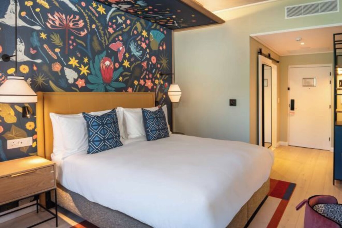 Une chambre d’hôtel avec un grand lit, une table de chevet, une lampe et une fresque murale aux motifs floraux et aux couleurs vives.