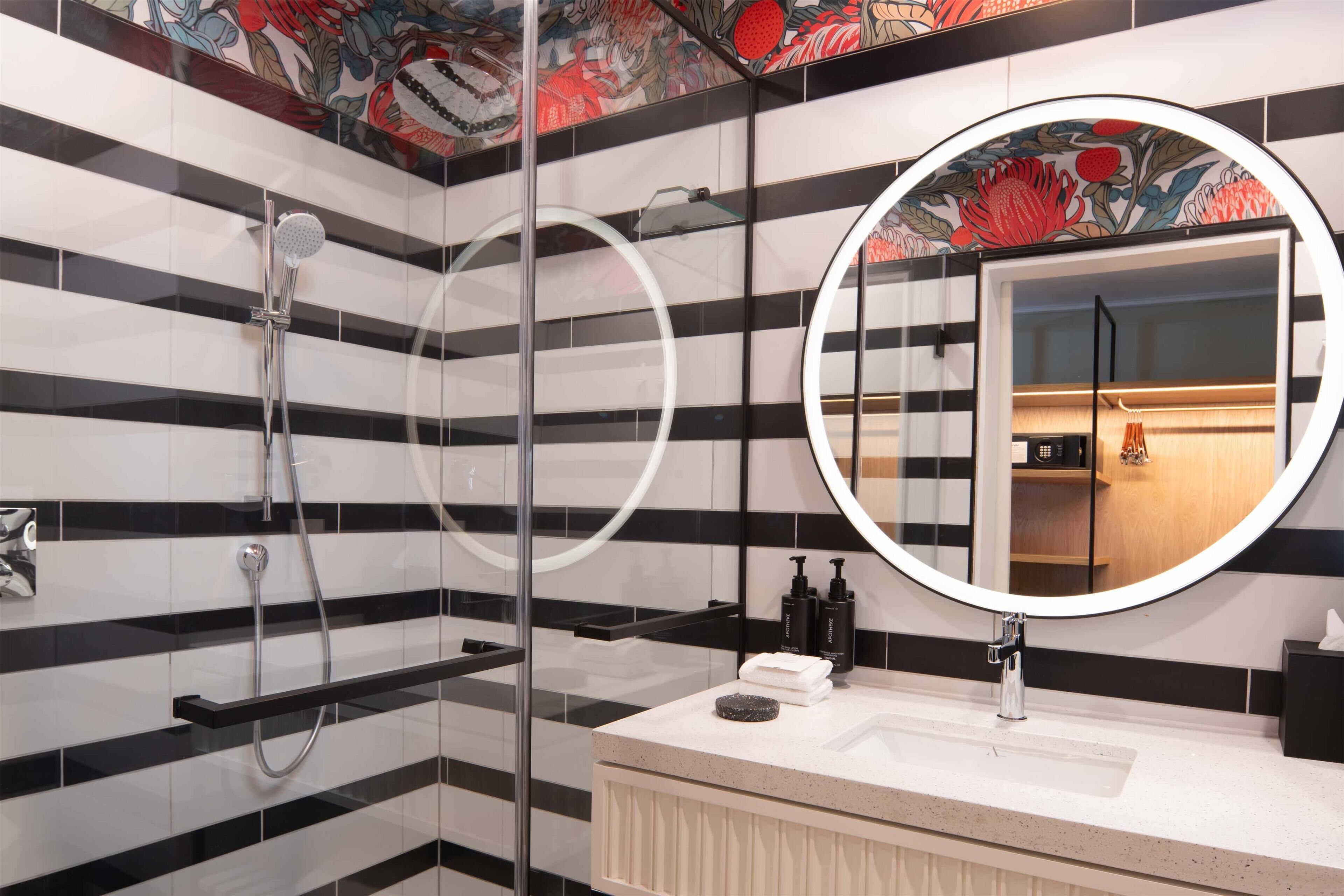 A modern bathroom with black and white striped tiles, a glass-enclosed shower, a round mirror, and a sink with toiletries.