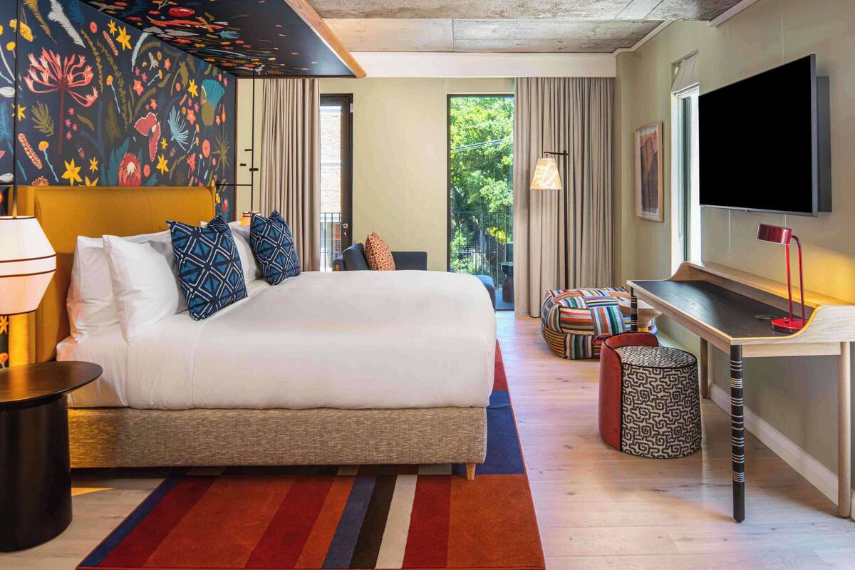 Una habitación de hotel moderna con una cama grande, un escritorio, un televisor y un balcón con vistas a zonas verdes.