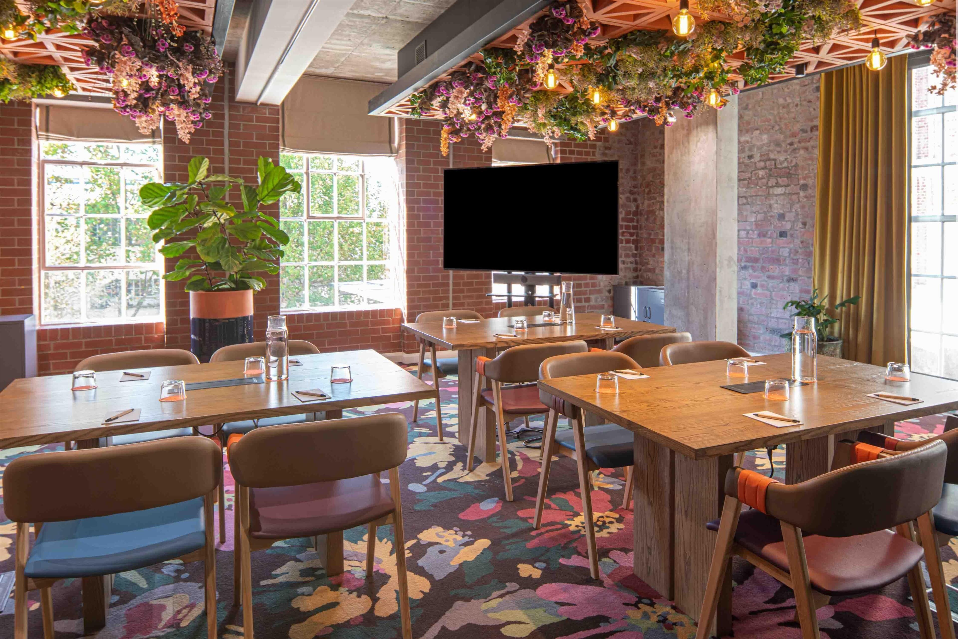 Une salle de conférence avec des tables en bois, des chaises colorées, une grande plante en pot, un téléviseur à écran plat et un mur en briques.