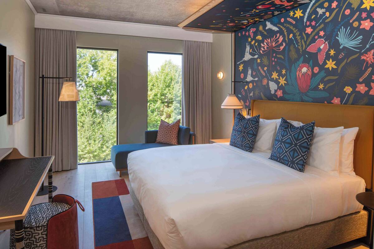 Habitación de hotel con una cama grande, un escritorio y una silla. Se aprecia una ventana con cortinas, una pared con un mural floral y un techo con un diseño texturizado.