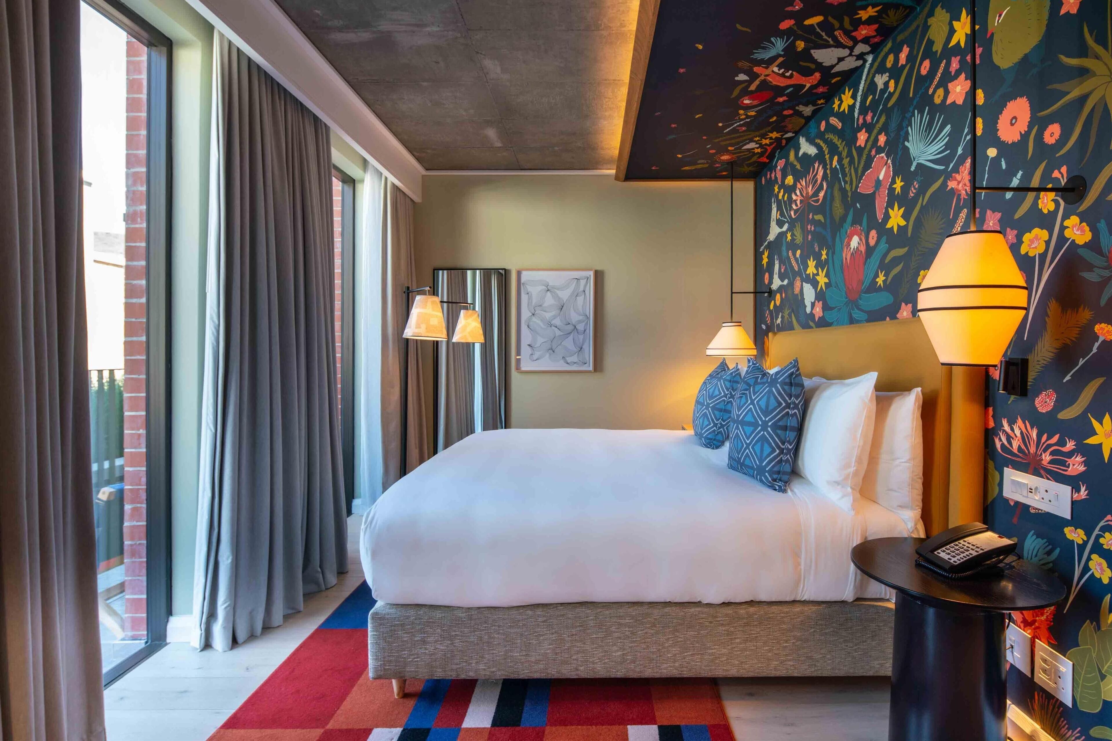 Une chambre d’hôtel avec un grand lit, une petite table de chevet avec un téléphone, un mur orné d’une fresque florale et une fenêtre avec des rideaux.