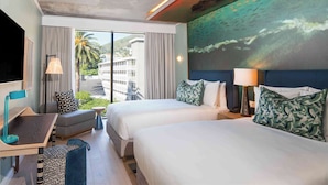 Habitación de hotel con dos camas, un escritorio, una silla y un ventanal con vistas a palmeras y edificios.