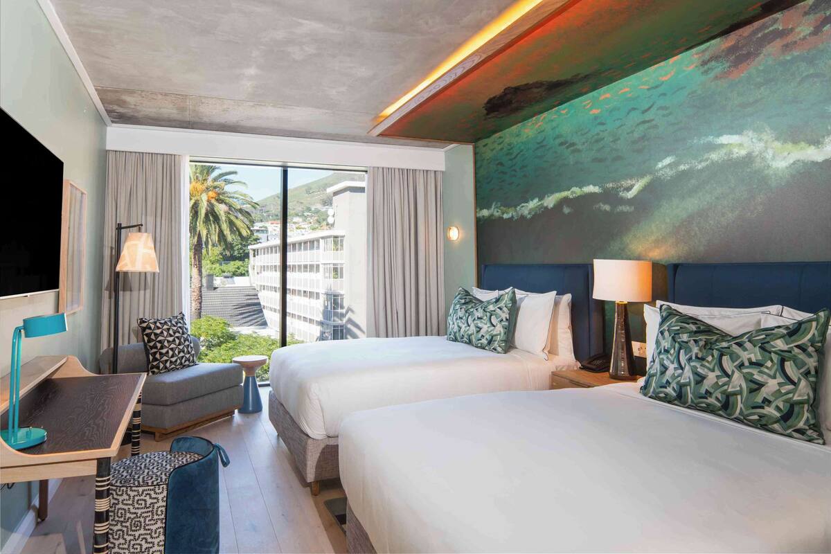 Habitación de hotel con dos camas, un escritorio, una silla y un ventanal con vistas a palmeras y edificios.