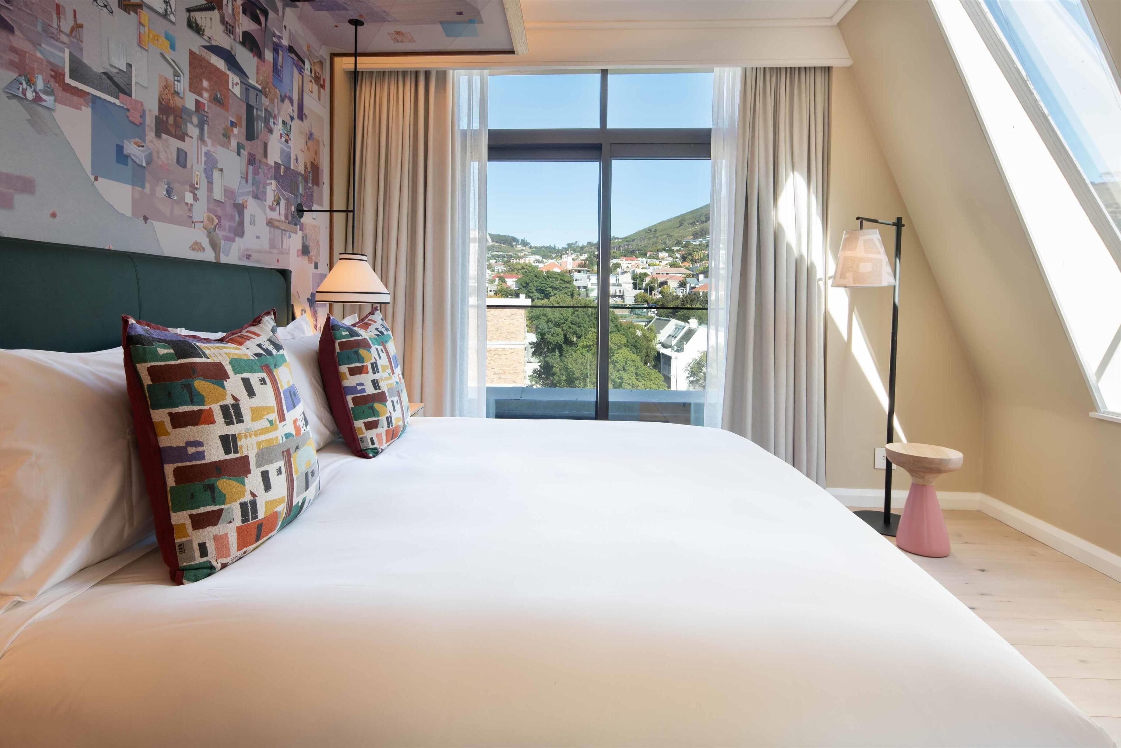 Ein Hotelzimmer mit einem großen Bett, farbenfrohen Kissen, Blick auf die Stadt und einer modernen Lampe.