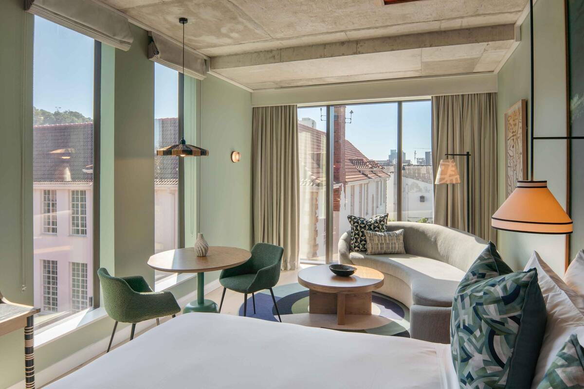 Habitación de hotel con cama, sofá, mesa redonda y una silla. Amplias ventanas con cortinas y vistas a la ciudad.