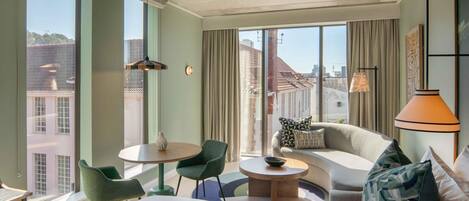 A hotel room with a bed, a sofa, a round table, and a chair. There are large windows with curtains, and a city view is visible outside.