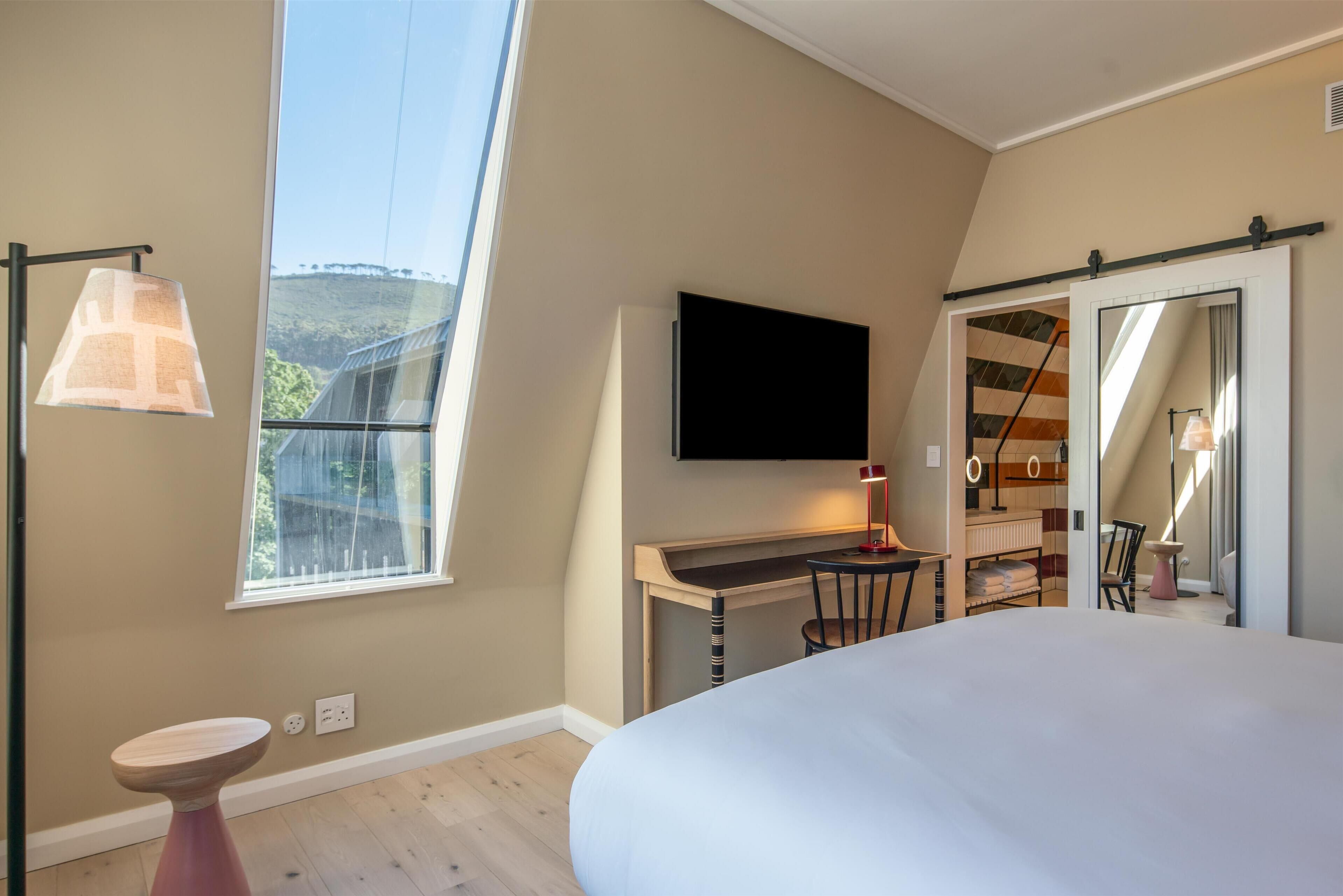 Un dormitorio con una cama grande, un escritorio con silla, un televisor montado en la pared y una ventana con vistas a una colina.