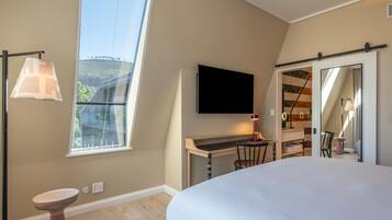 Un dormitorio con una cama grande, un escritorio con silla, un televisor montado en la pared y una ventana con vistas a una colina.