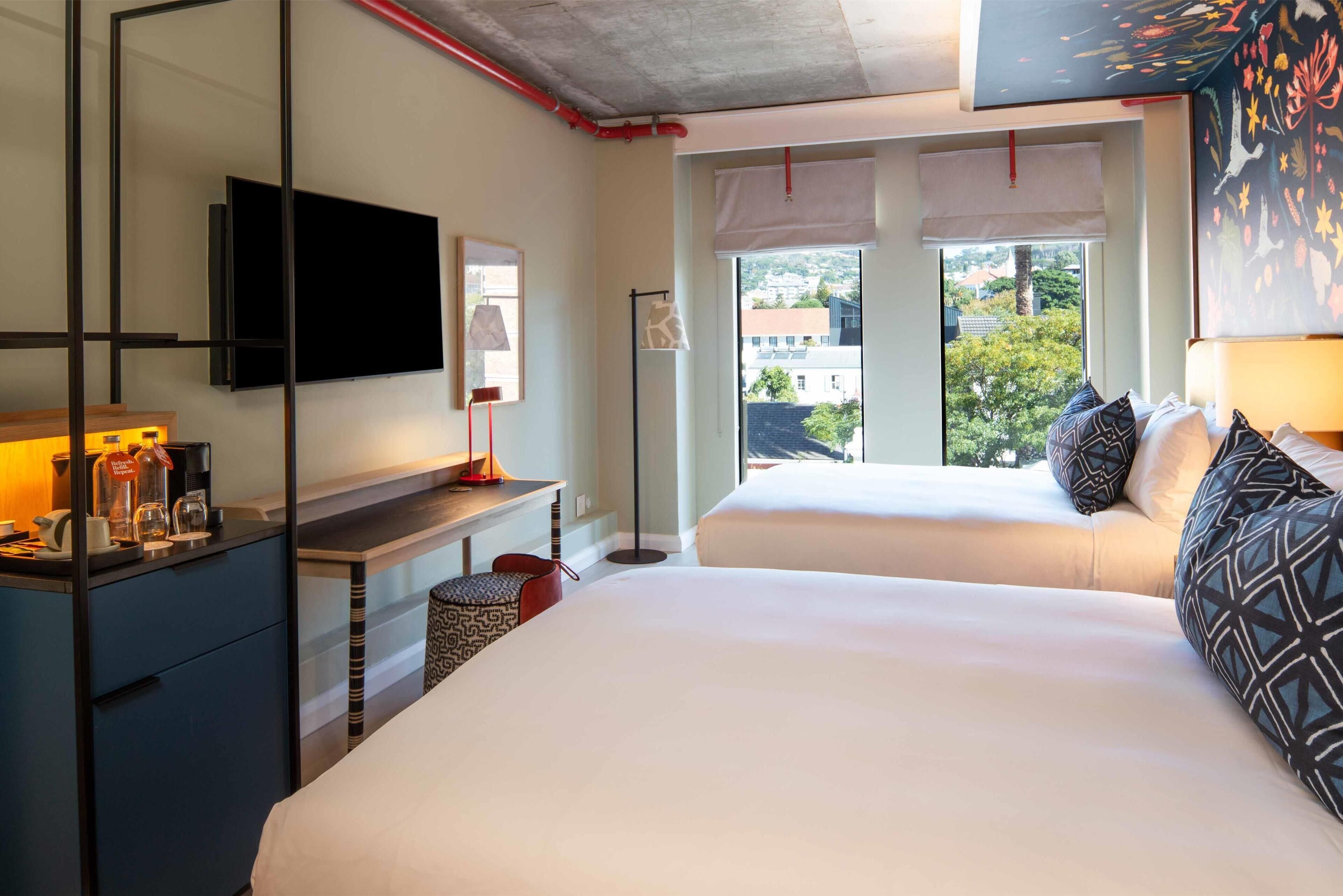 Une chambre d’hôtel avec un grand lit, un bureau doté d’un téléviseur à écran plat, une fenêtre donnant sur des immeubles et un plafond avec des tuyaux apparents.