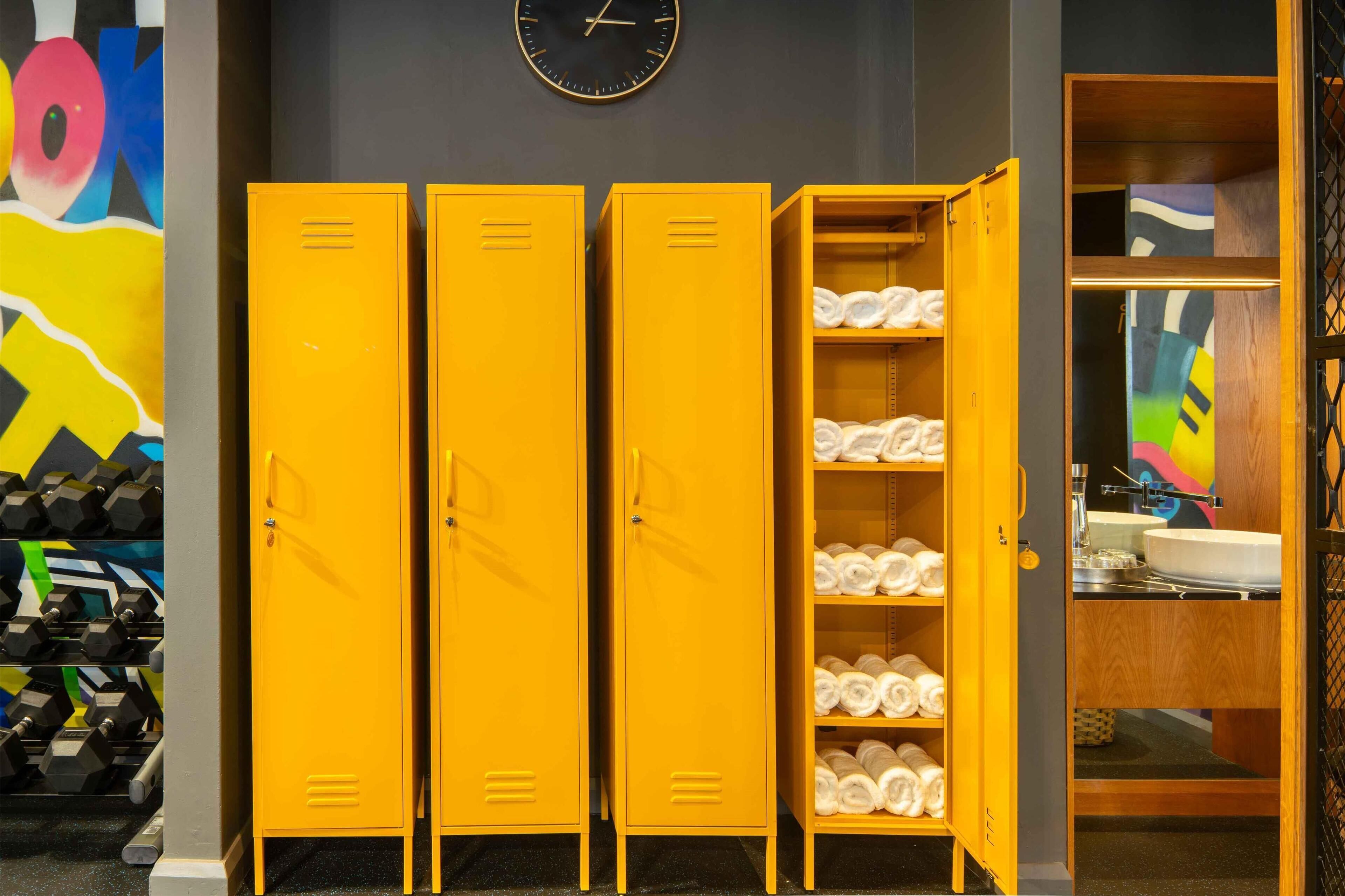 A set of yellow lockers with towels inside, a clock on the wall, and a colorful abstract painting.