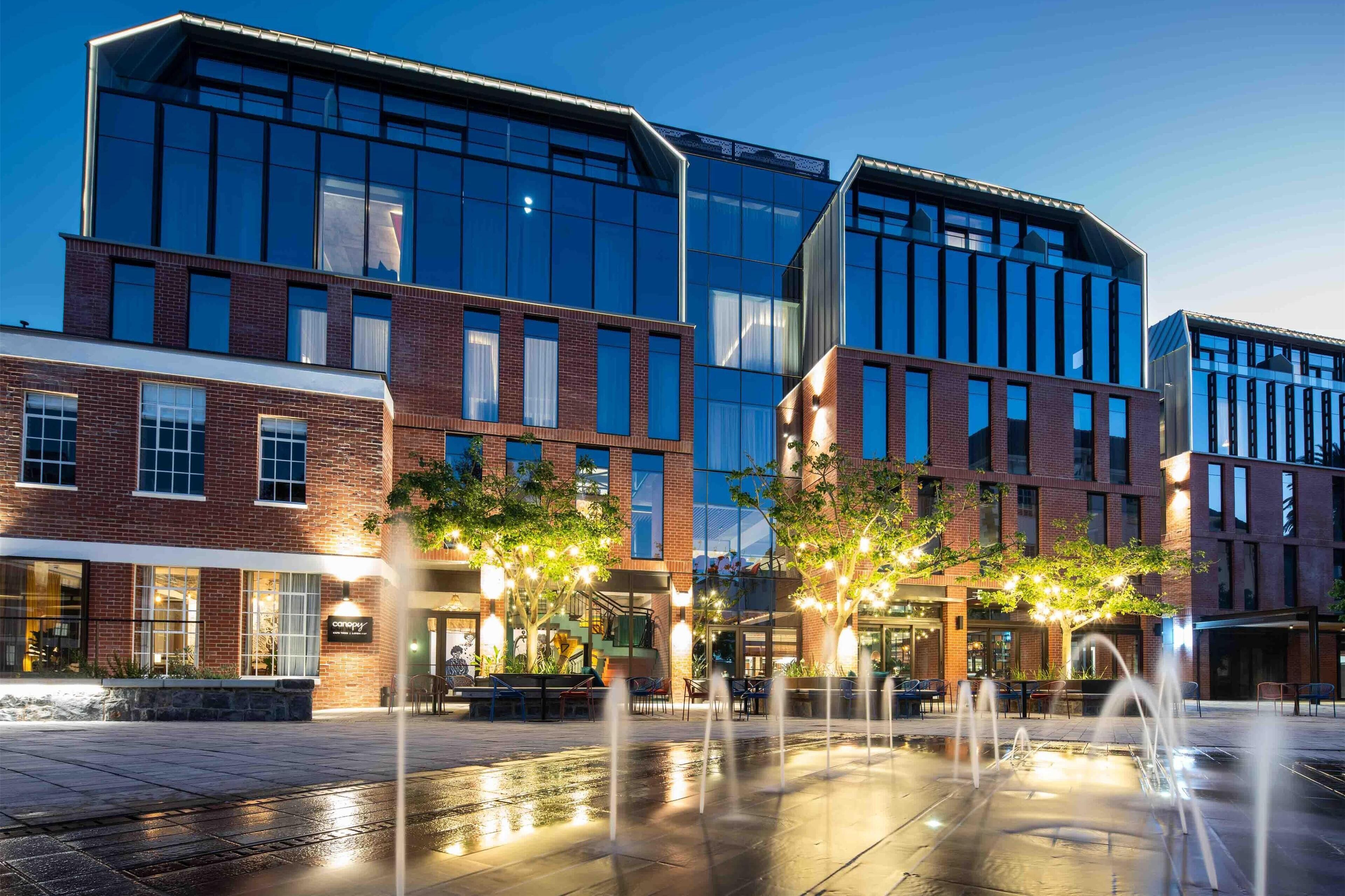 A modern building with a brick facade and large windows, a courtyard with a fountain, and outdoor seating.