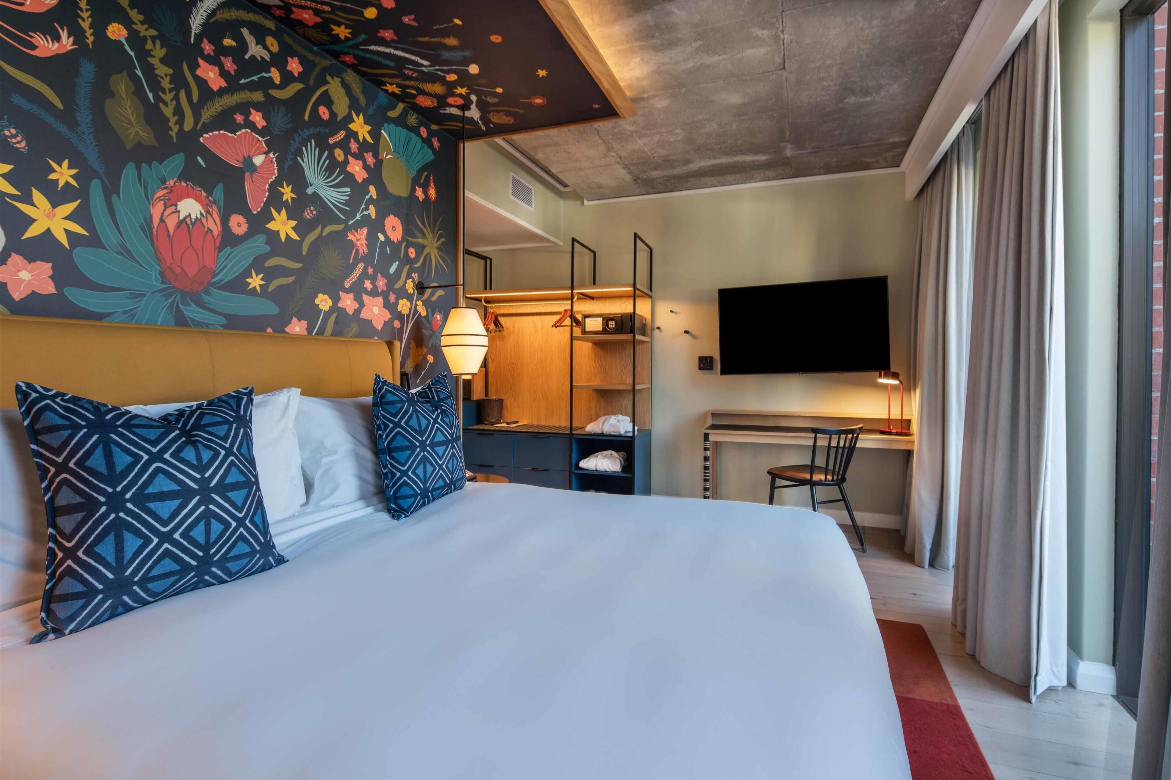A hotel room with a large bed, a desk, a TV, and a colorful floral mural on the wall.