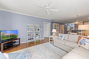 A living room with a television, a white shelving unit, a sofa with decorative pillows, and a ceiling fan. - Mini Weeks Stays Available Sunset Island 4 Fountai (Ocean City)