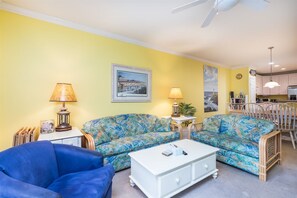 Ein Wohnzimmer mit einer Couch, einem Couchtisch und einem blauen Sessel. Im Hintergrund befindet sich ein Küchenbereich.